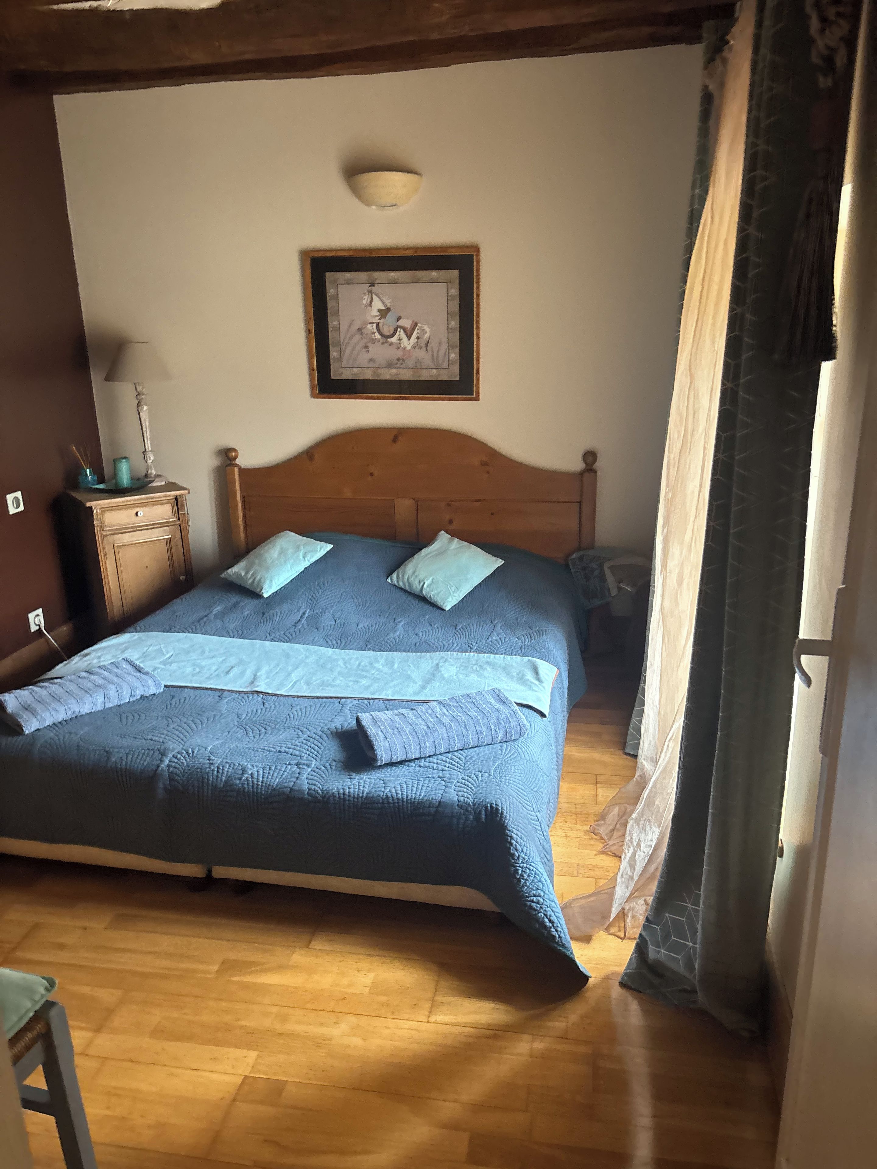 Quarto com cama de madeira, mesa de cabeceira, luminária, quadro na parede e janela com cortinas.