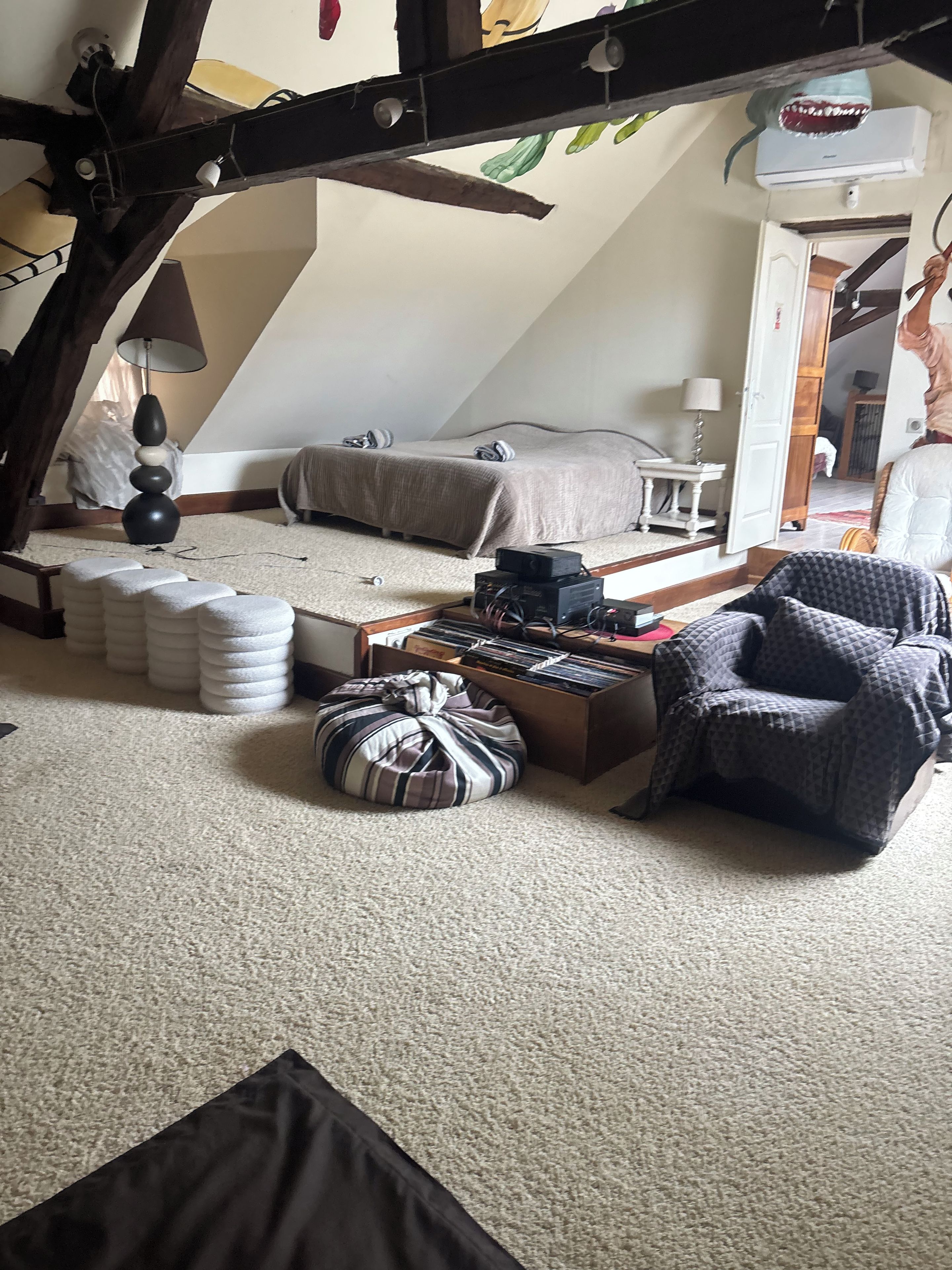 A loft bedroom with a sloped ceiling, a bed, a desk with electronic equipment, a chair, and a bean bag.