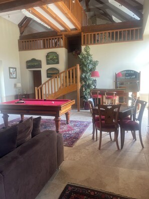 A spacious living area with a pool table, a sofa, and a dining table with chairs. A wooden staircase leads to an upper level.