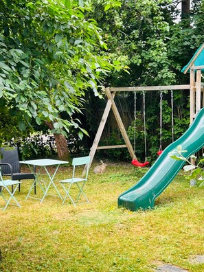 A wooden swing set with a green slide, a small table, and two chairs in a grassy outdoor area.