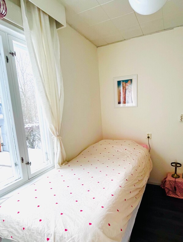 Una habitación con cama, ventana con cortinas, un cuadro en la pared y una mesita de noche con lámpara.