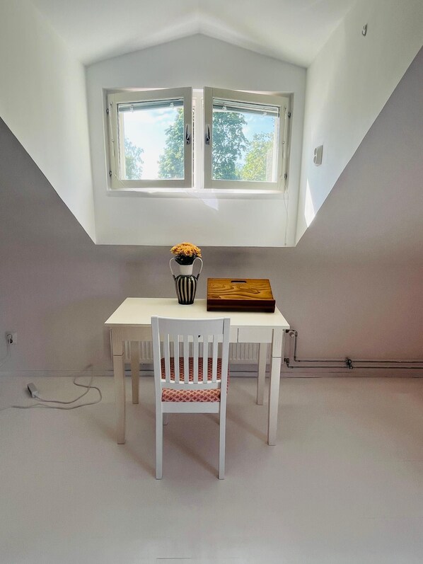 A room with a white table and chair, a window with a view of trees, and a power outlet on the wall.