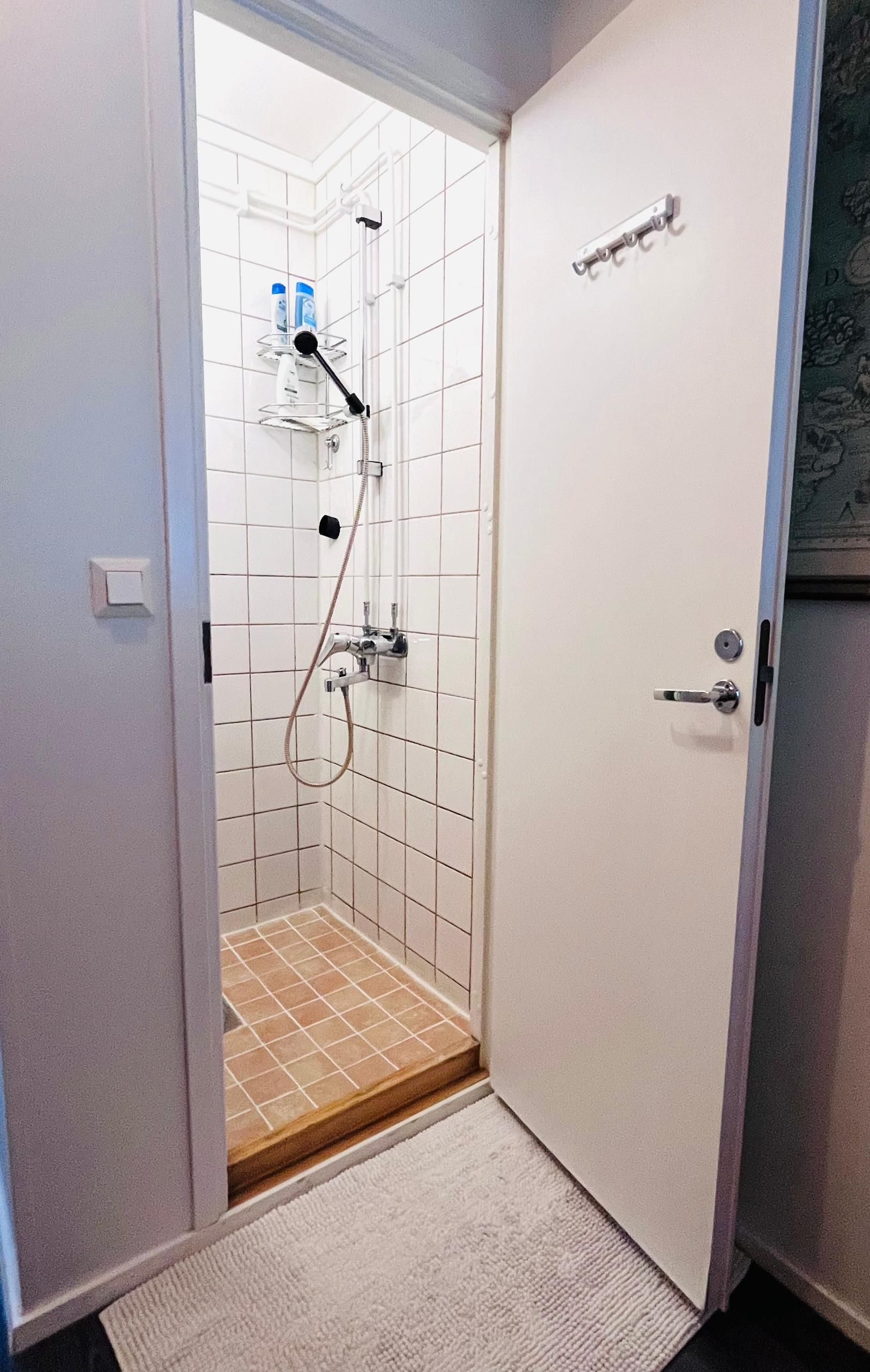 A bathroom with white tiled walls, a shower with a handheld showerhead, and a door with a handle.