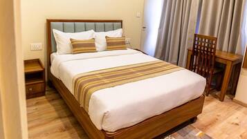 Una cama bien tendida con estructura de madera y cubrecama blanco, una mesita de noche de madera con lámpara, y una silla con estructura de madera.