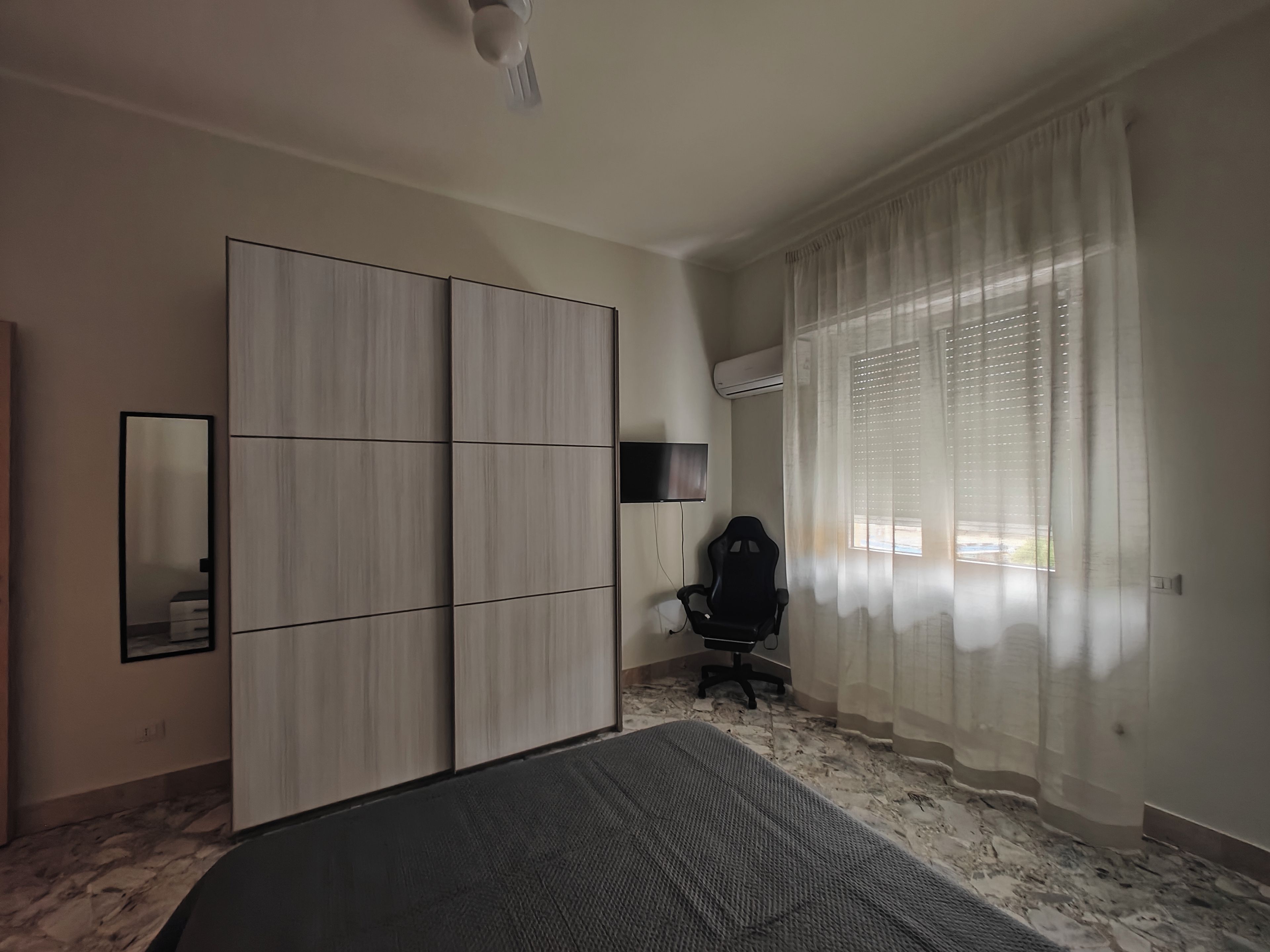 A bedroom with a bed, a wardrobe, a desk, a chair, a TV, and a window with curtains.
