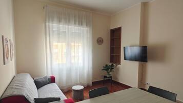 A room with a dining table, a sofa, a ceiling fan, a TV mounted on the wall, and a window with curtains.