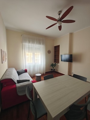 Una habitación con una mesa de comedor, un sofá, un ventilador de techo, un televisor montado en la pared y una ventana con cortinas.