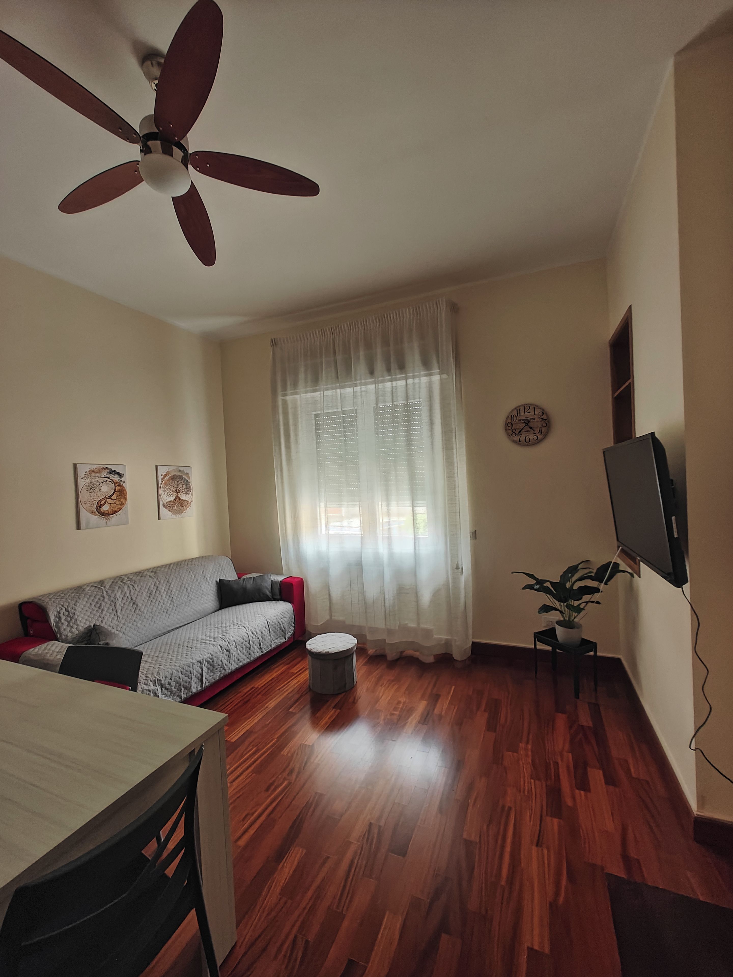 Un salon avec un ventilateur de plafond, un canapé, une table à manger, un petit pouf rond, une plante en pot et une horloge murale.