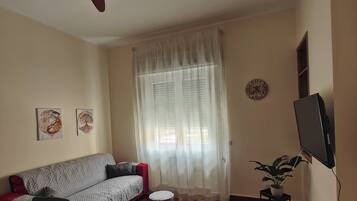 Un salon avec un ventilateur de plafond, un canapé, une table à manger, un petit pouf rond, une plante en pot et une horloge murale.