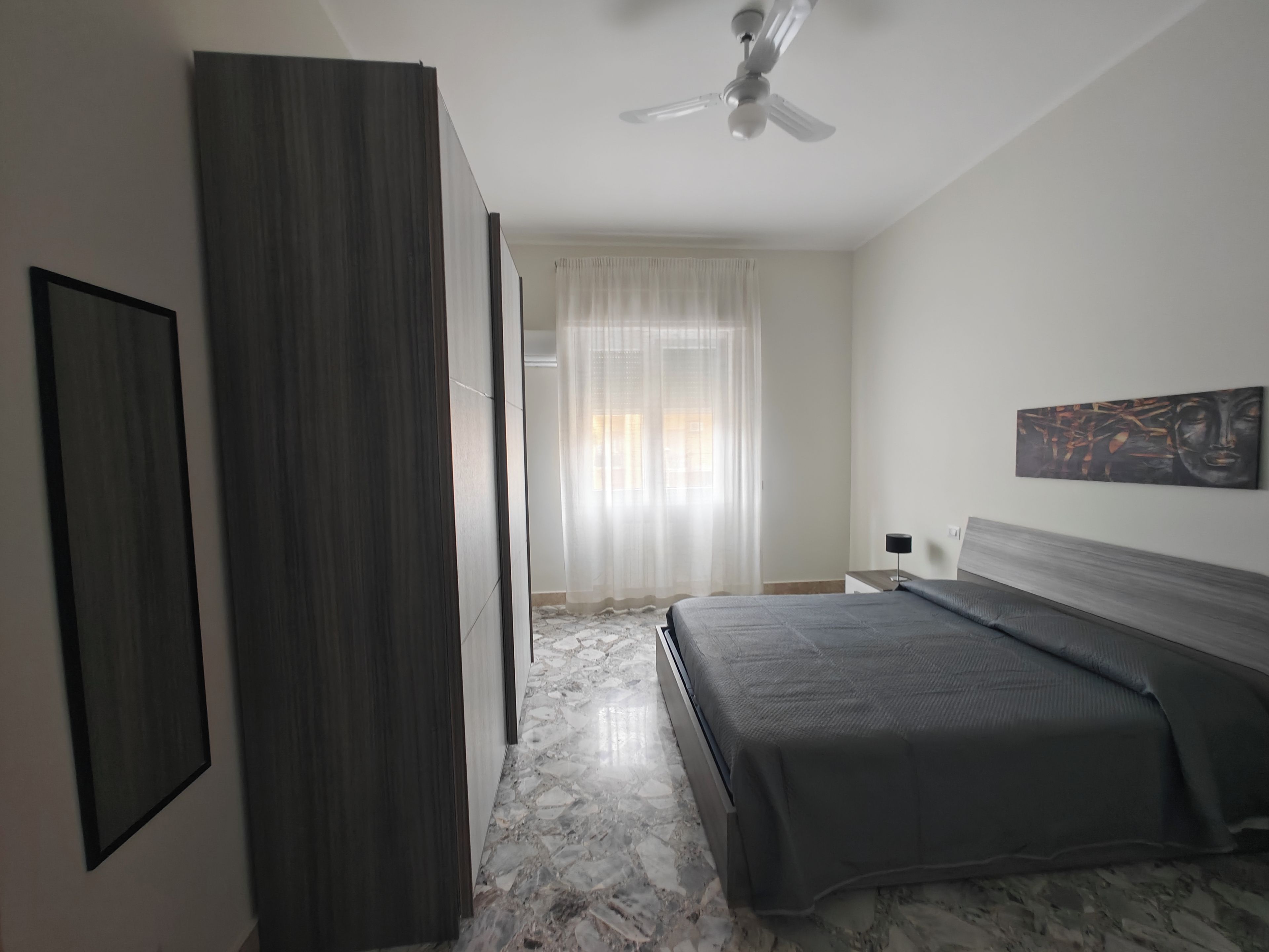 Een slaapkamer met een bed, een plafondventilator, een raam met gordijnen en een schilderij aan de muur.
