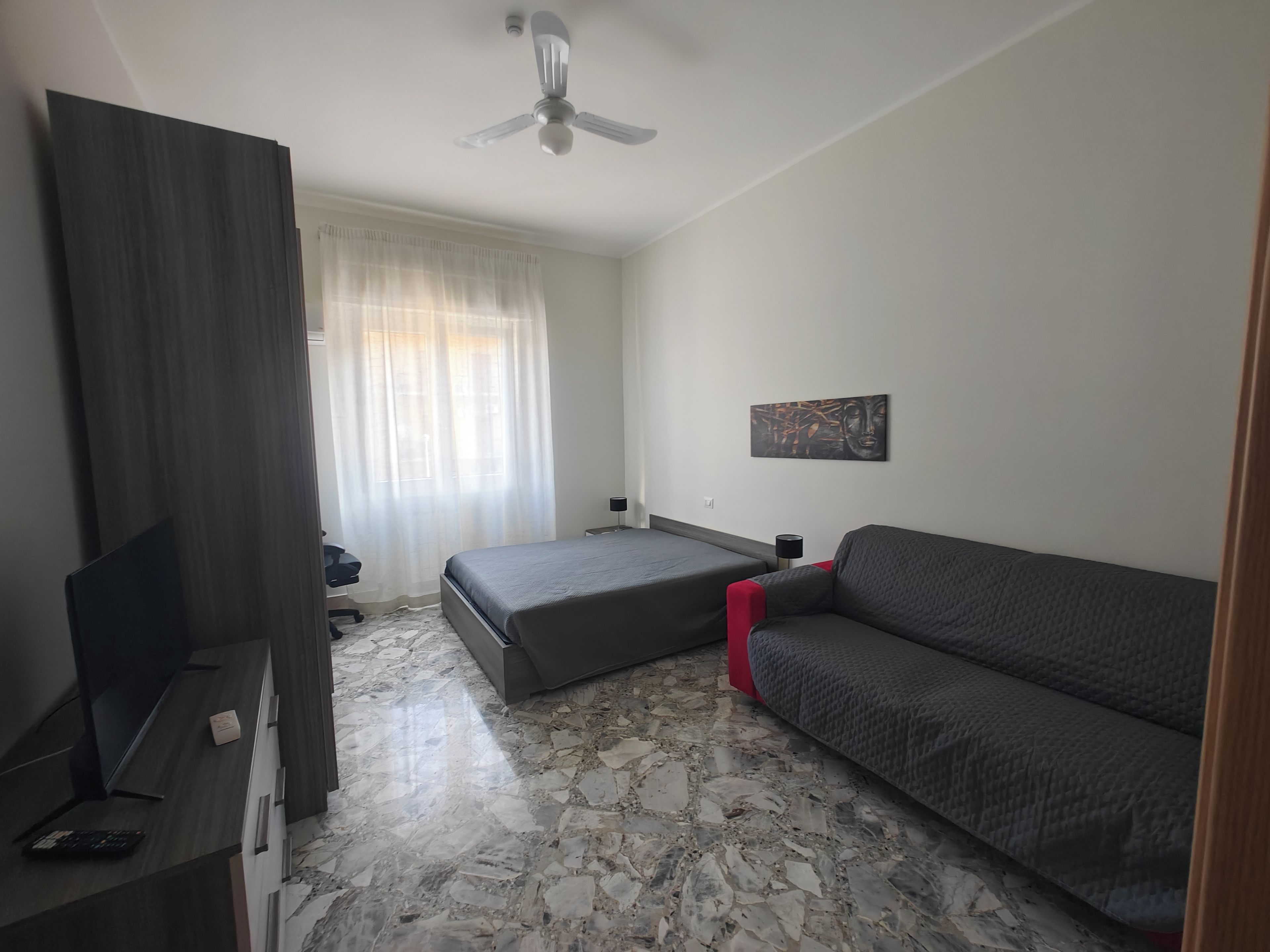 A bedroom with a bed, a sofa, a desk, a chair, a ceiling fan, and a window with curtains.