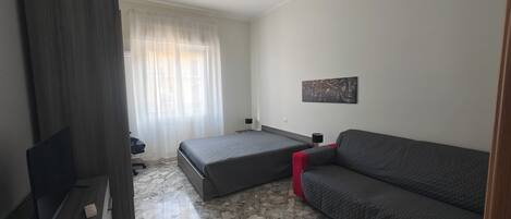 A bedroom with a bed, a sofa, a desk, a chair, a ceiling fan, and a window with curtains.
