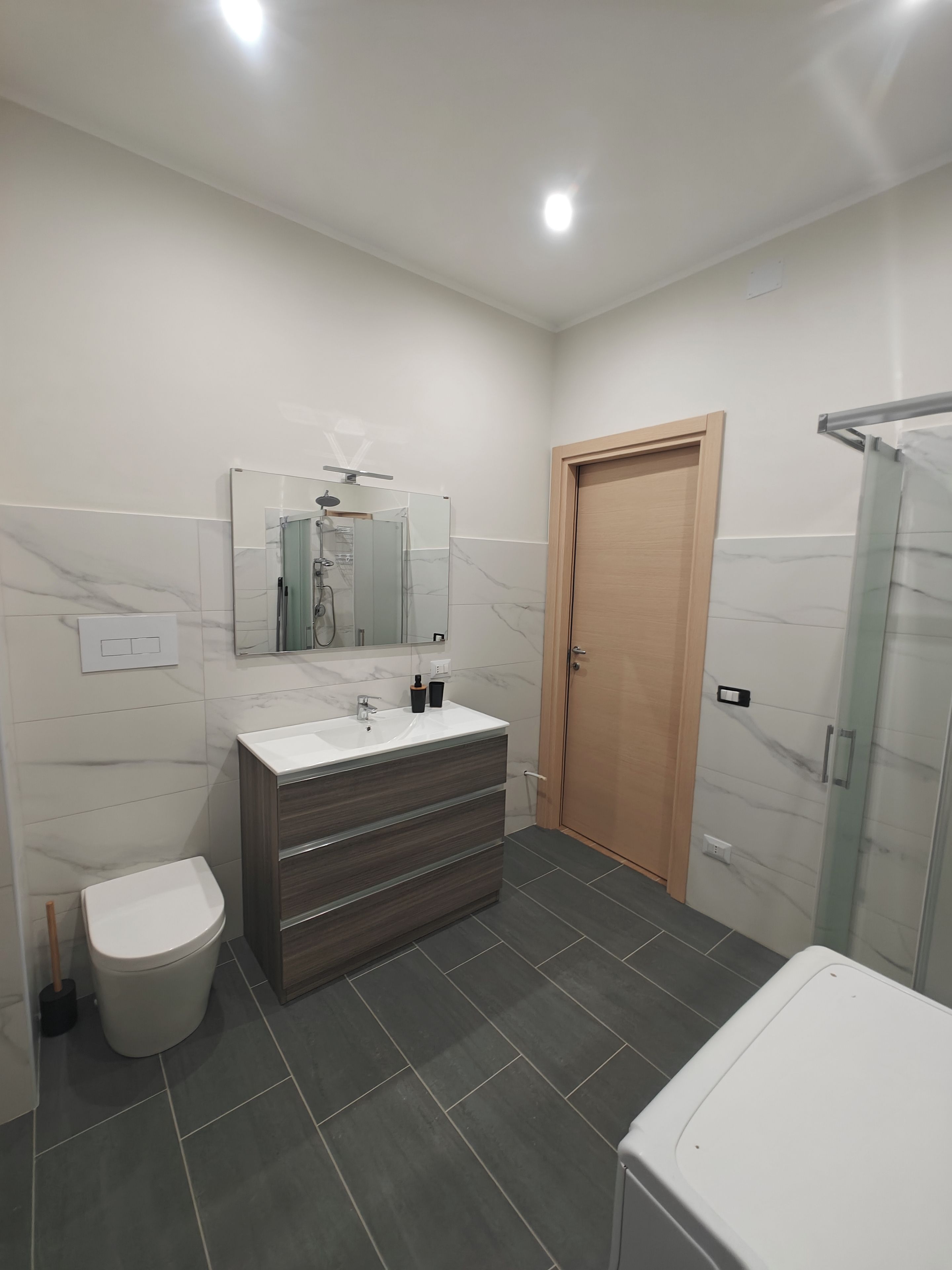 Une salle de bain moderne avec un lavabo blanc, un meuble-lavabo en bois foncé, une cabine de douche en verre et une porte en bois.