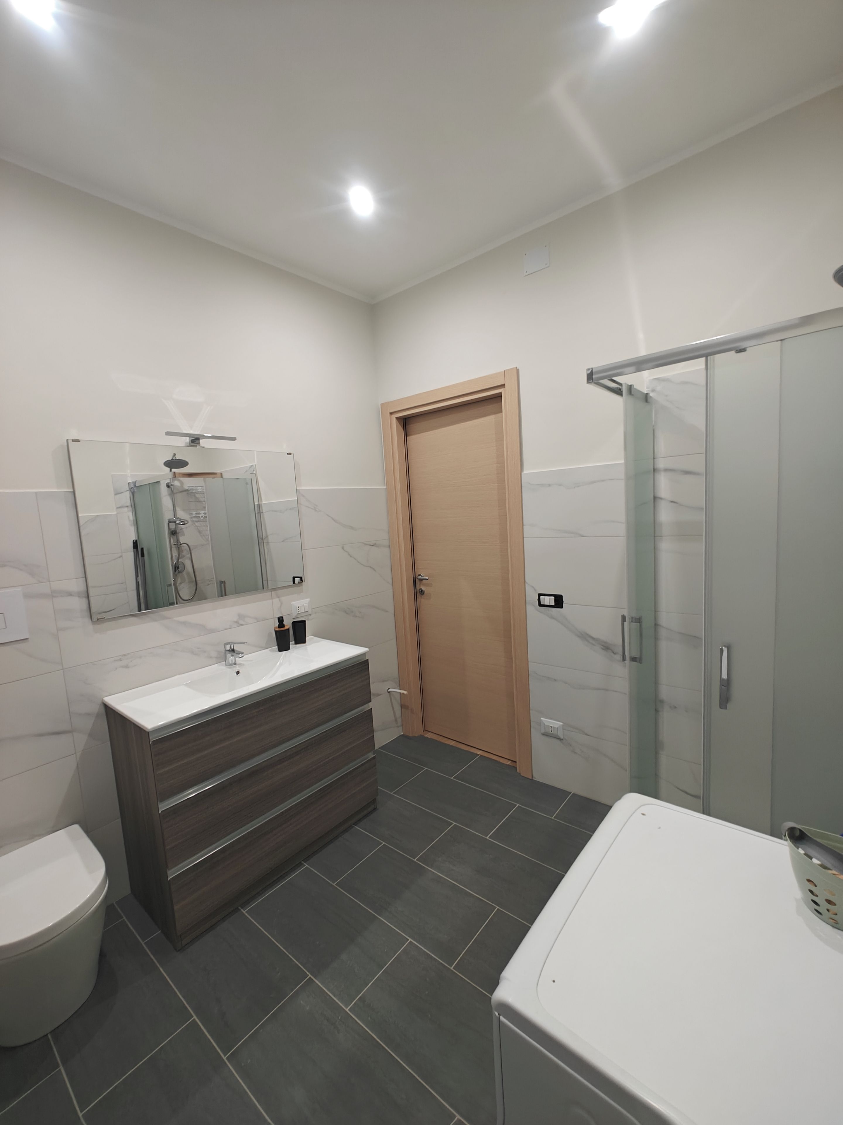 A modern bathroom with a white sink, dark wooden vanity, a glass shower enclosure, and a wooden door.