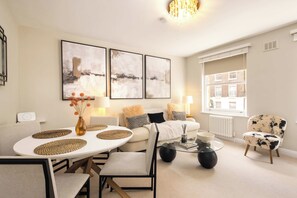 Ein modernes Wohnzimmer mit einem weißen Sofa, einem runden Couchtisch und einem Esstisch mit gewebten Unterlagen. An der Wand hängen abstrakte Gemälde, eine Chandelier und ein Fenster mit Blick auf Gebäude.