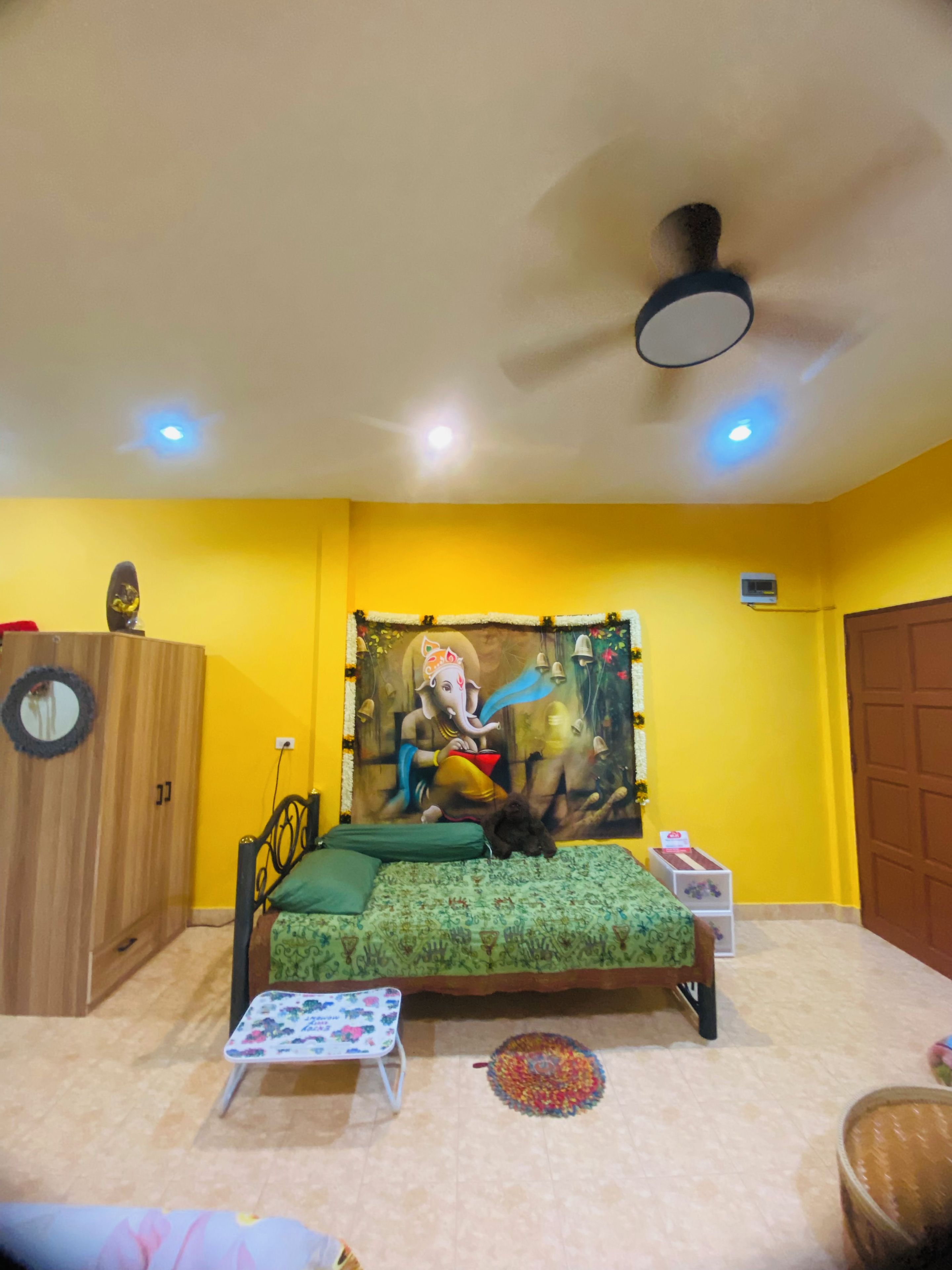 A room with a bed, a small table, a chair, a ceiling fan, and a wall tapestry.