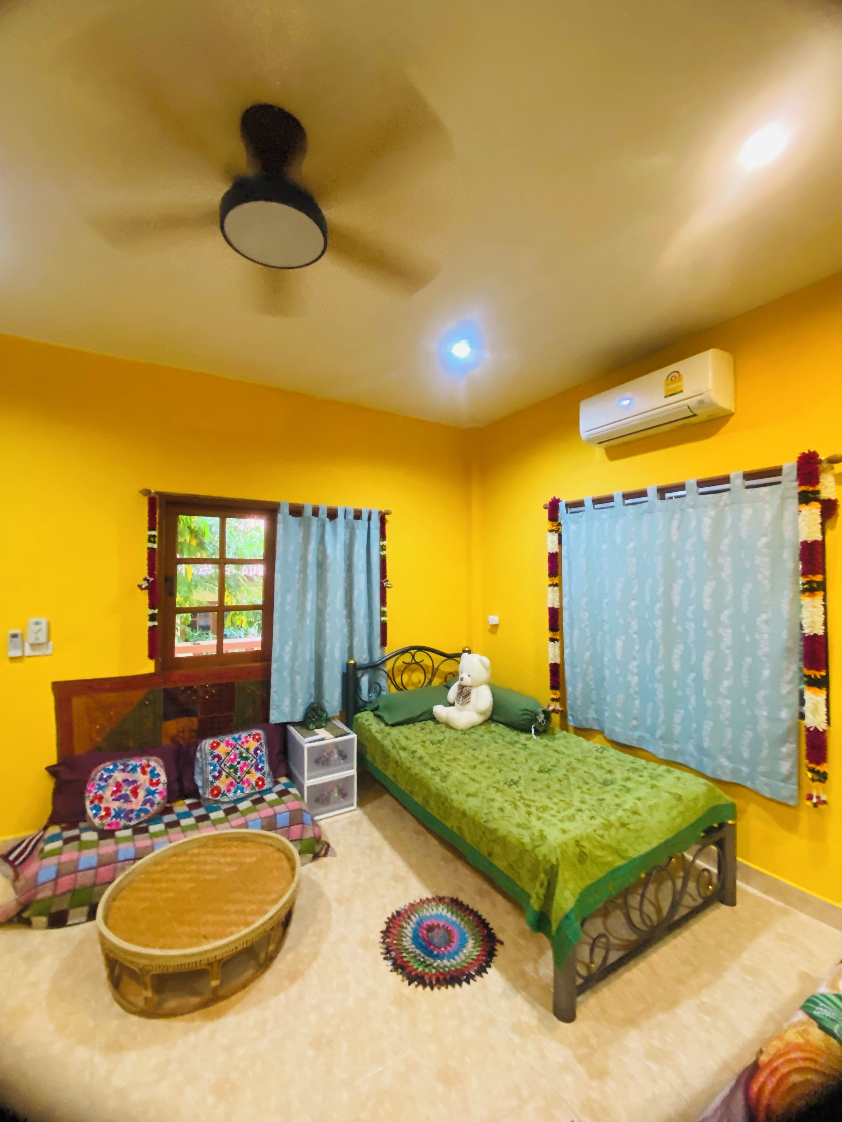 A room with a bed, a window with curtains, a ceiling fan, an air conditioning unit, and a small table with a chair.