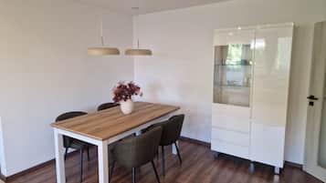Ein Essbereich mit einem Holztisch, vier Stühlen, einer Blumenvase, Pendelleuchten und einem weißen Schrank mit Glastüren.