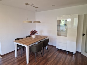 A dining area with a wooden table, four chairs, a vase with flowers, pendant lights, and a white cabinet with glass doors.
