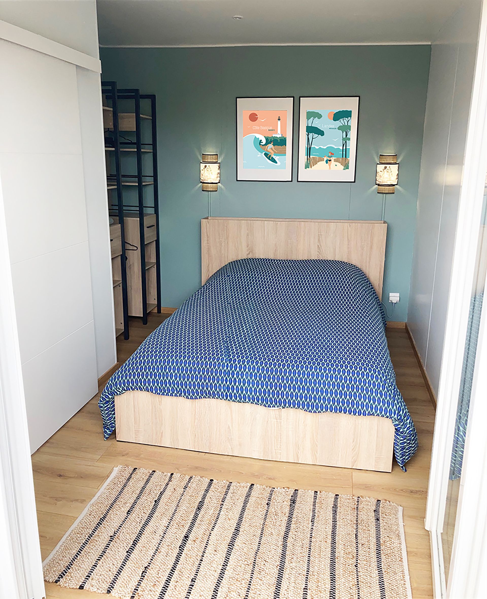 A bedroom with a bed, a wooden headboard, two wall-mounted lamps, a framed picture, a woven rug, and a ladder against the wall.