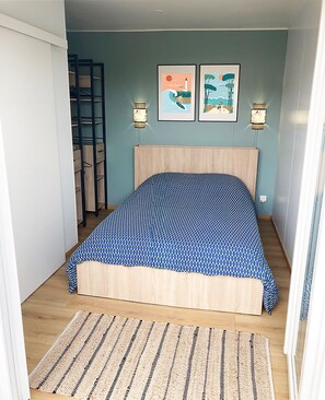 Un dormitorio con cama, cabecera de madera, dos lámparas fijadas a la pared, un cuadro enmarco, alfombra tejida y una escalera apoyada en la pared.