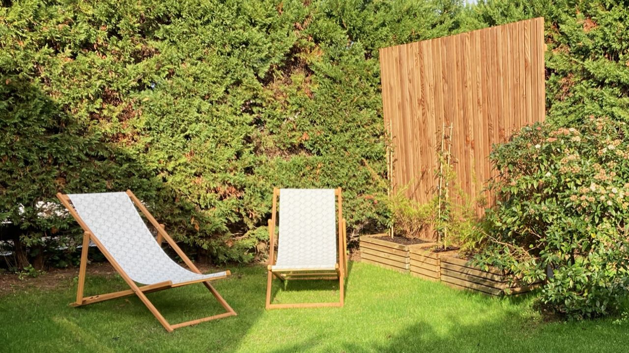 Dos sillas de madera con cojines, una valla de madera y un jardín bien cuidado con diversos arbustos.