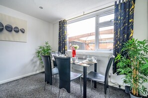 Un comedor con una mesa dispuesta para cuatro, sillas, una planta en maceta y una ventana con cortinas.
