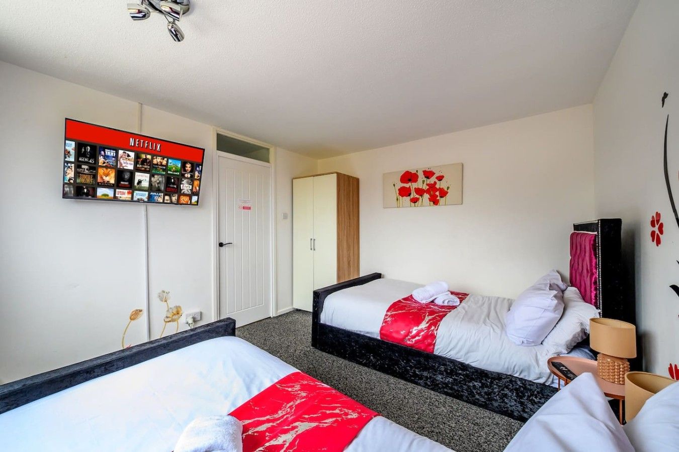 A hotel room with two beds, a wall-mounted TV displaying Netflix content, a door, and a small table with a lamp.