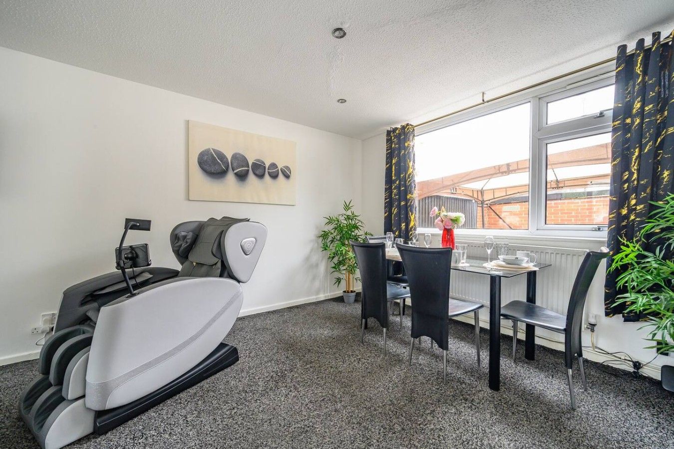 A room with a massage chair, a dining table with chairs, a window with curtains, and a painting on the wall.