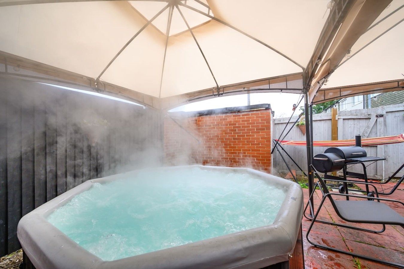 A hot tub under a canopy with a brick wall and a barbecue grill in the background.