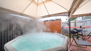 A hot tub under a canopy with a brick wall and a barbecue grill in the background.