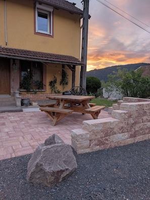 Ein gelbes Gebäude mit einer Terrassenfläche, die einen Holztisch und Bänke bietet, umgeben von Steinstufen und einem großen Felsen.