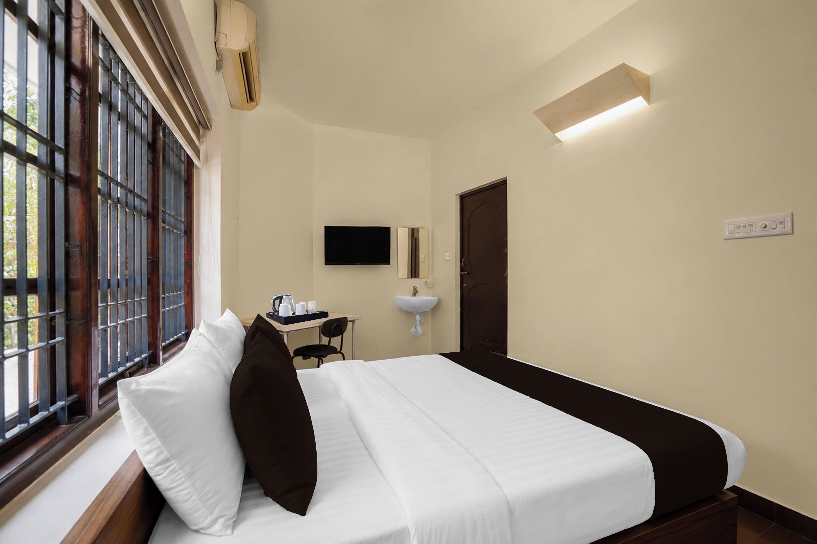 A hotel room with a bed, a desk with a chair, a TV, a sink, and a window with bars.