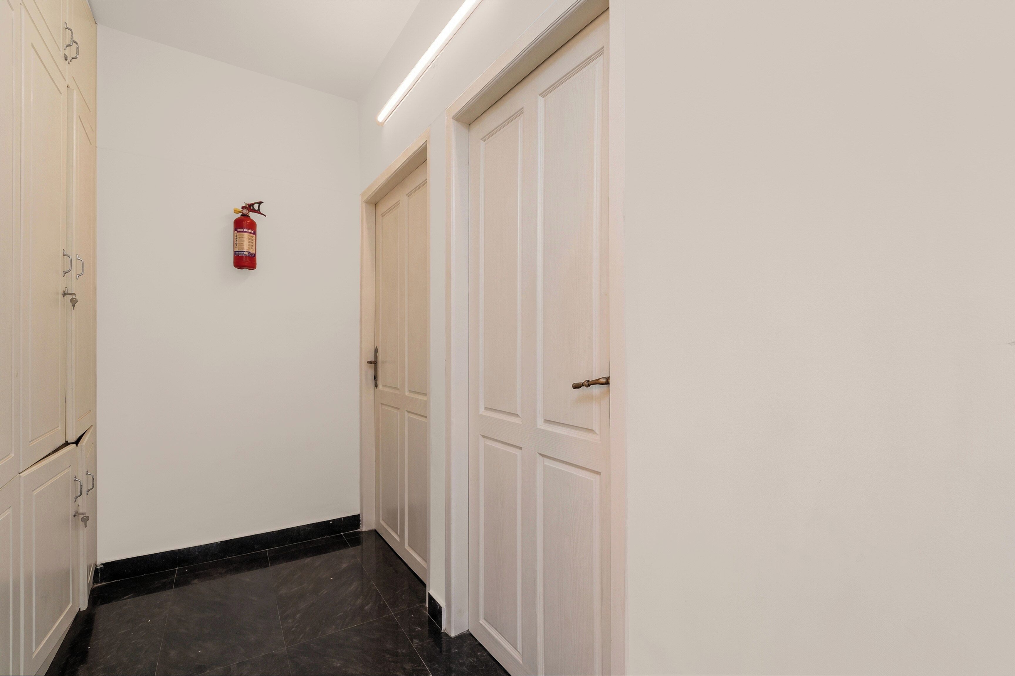 Un couloir avec des portes blanches, un extincteur fixé au mur et un sol recouvert de carreaux noirs.