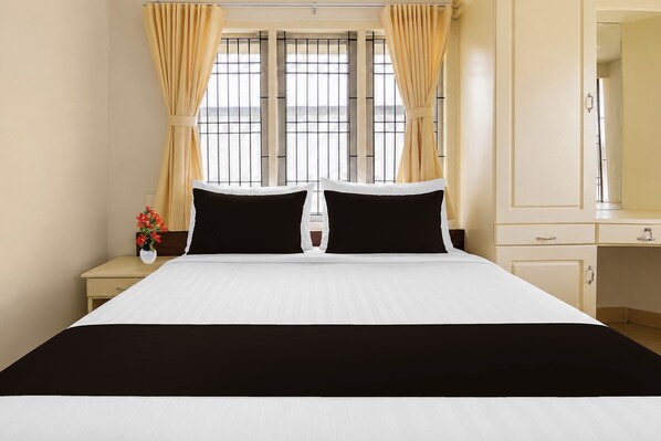 A hotel room with a large bed, two black pillows, a white bedspread, a nightstand with a vase of flowers, and a window with curtains.