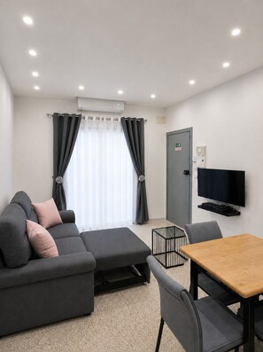 A modern living room with a sectional sofa, a dining table, and a flat-screen TV mounted on the wall. - 2-Bedroom Modern Apartment in Xlendi,Gozo (Il-Munxar)