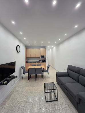 Ein modernes Wohnzimmer mit Flachbildfernseher, grauem Sofa, Esstisch mit Stühlen, Küchenzeile mit Holzschränken und Fliesenboden.