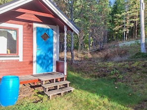 Eine kleine Holzhütte mit blauer Tür, einer Holzterrasse und einem bewaldeten Hintergrund.
