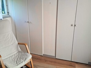 Una habitación con una silla blanca, un armario de dos puertas y una ventana con vista a la vegetación.