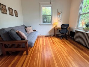Een kamer met een houten vloer, een grijze bank met gestreepte kussens, een zwarte bureaustoel, een kleine tafel met een lamp en een radiator.