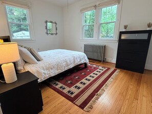 Een slaapkamer met een bed, een radiator, een dressoir en een raam met uitzicht op bomen.