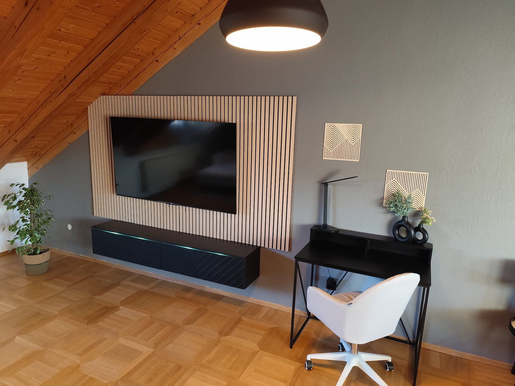 Una sala de estar moderna con un televisor de pantalla plana grande montado en una pared de paneles de madera, un escritorio negro con una silla de oficina blanca y una planta en maceta.