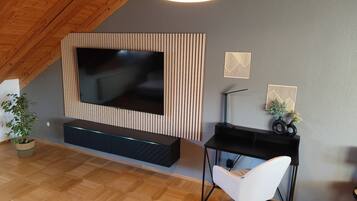 Una sala de estar moderna con un televisor de pantalla plana grande montado en una pared de paneles de madera, un escritorio negro con una silla de oficina blanca y una planta en maceta.