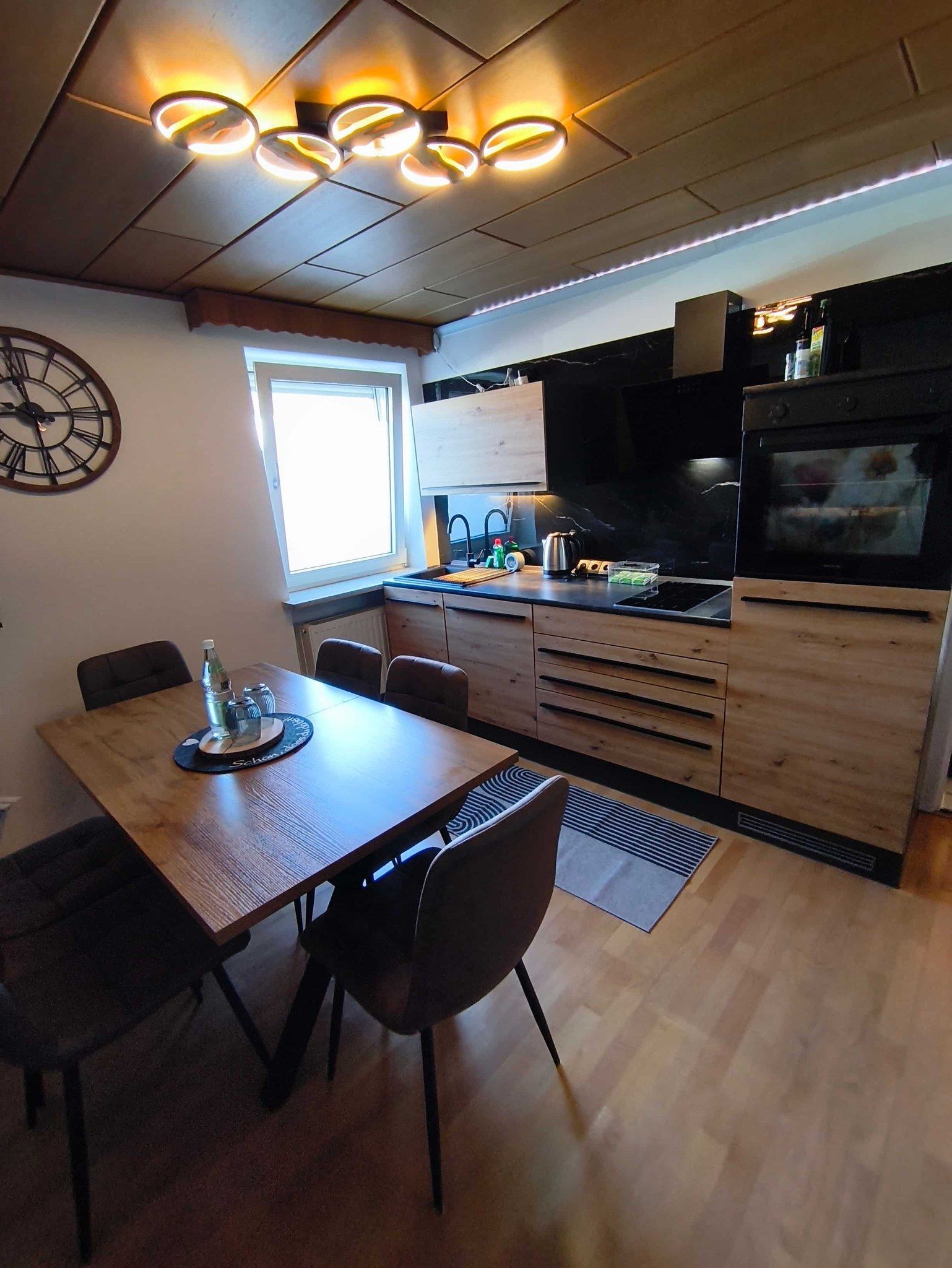 Una cocina moderna con zona de comedor, armarios de madera, un reloj grande y una ventana.