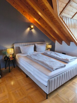 Un dormitorio con techo inclinado, una cama con ropa de cama blanca, dos lámparas de noche y una mesita con lámpara.