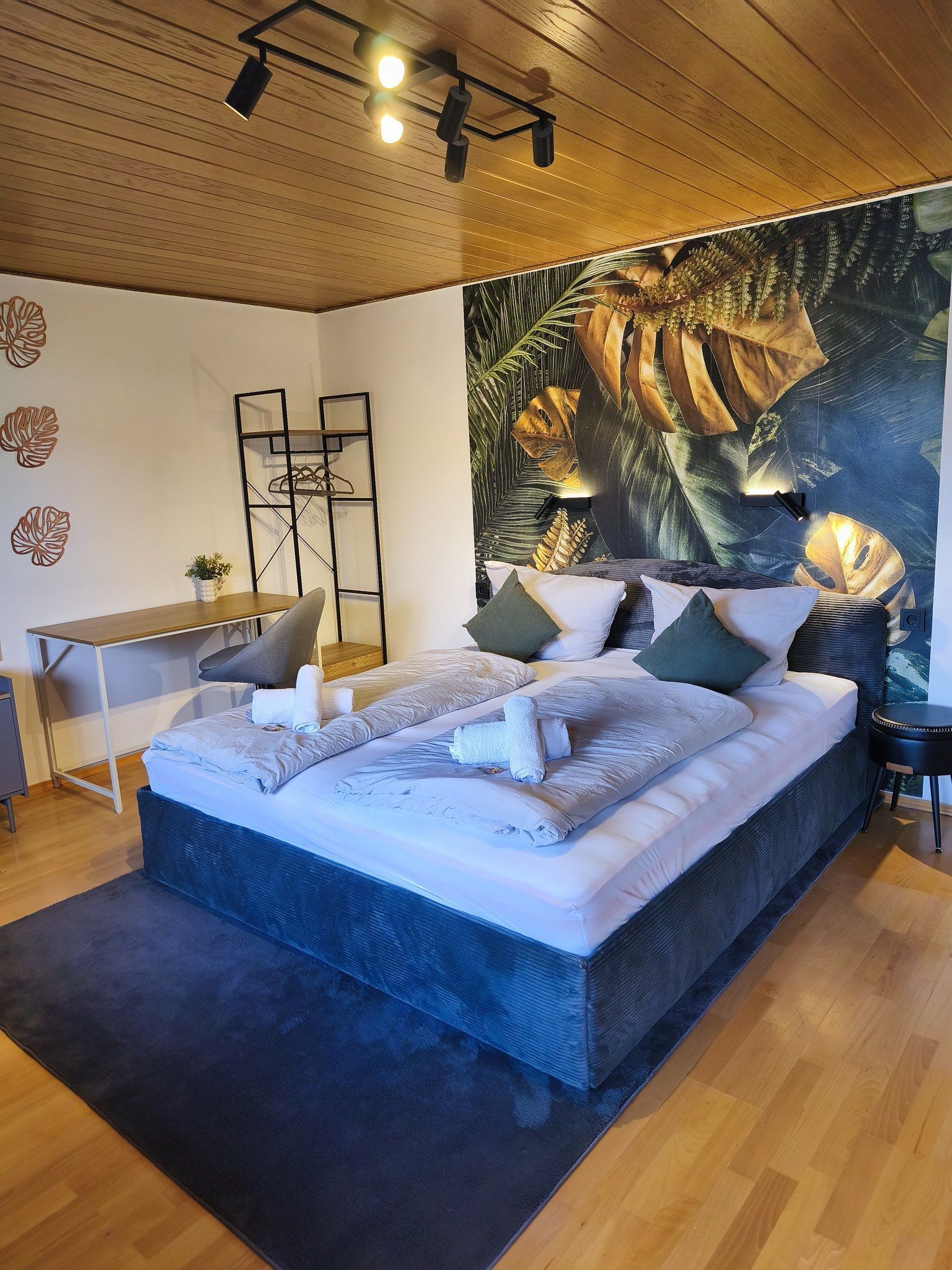 A modern bedroom with a large bed, a desk, and a tropical-themed wall mural.