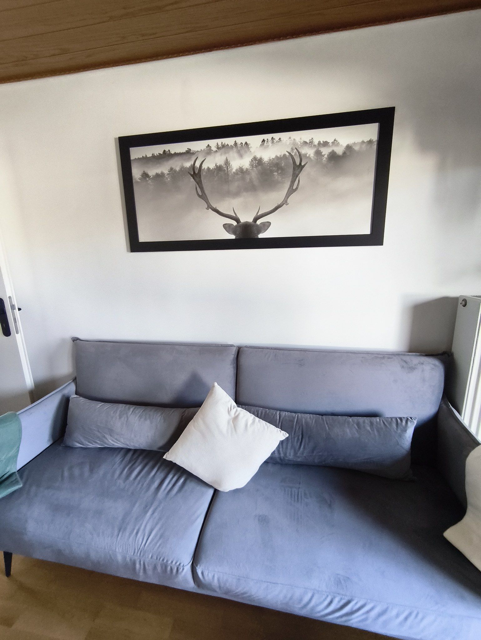Een grijze bank met een wit kussen, een lijst met een foto van een hert en een houten plafond.