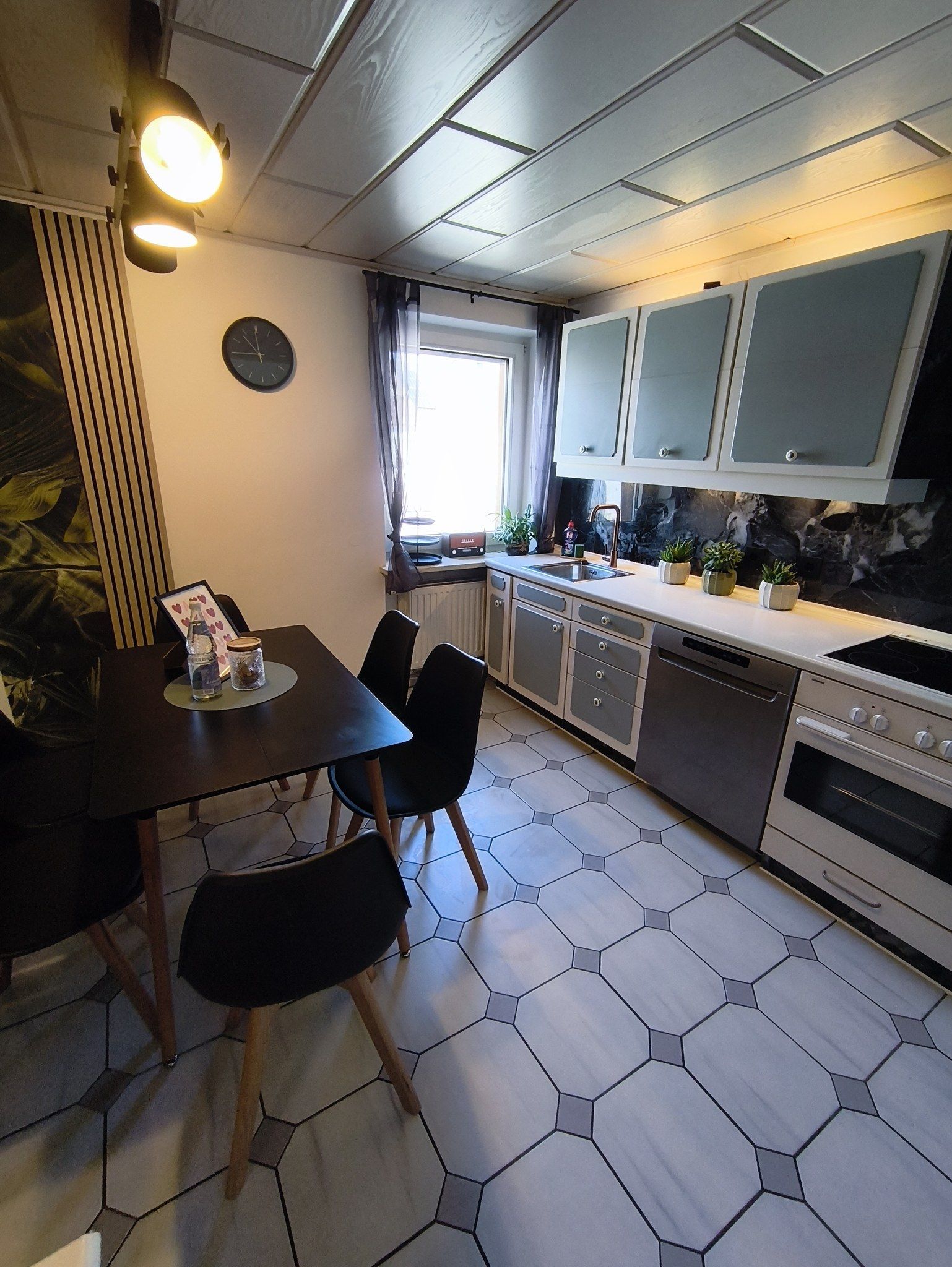 A modern kitchen with a dining area, featuring hexagonal tiles, a built-in oven, and a window with curtains.
