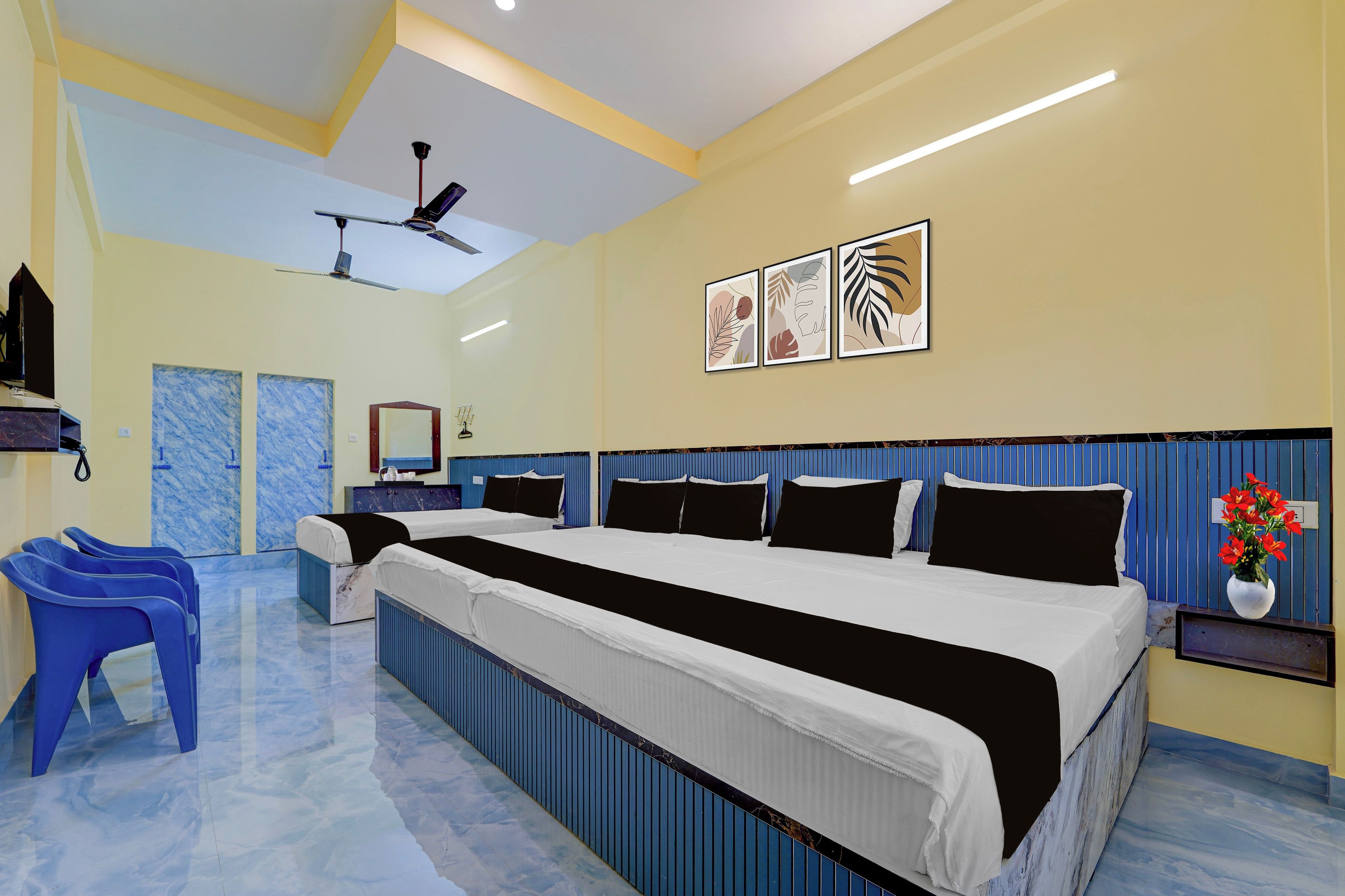 Quarto de hotel com uma cama grande, um ventilador de teto, televisão fixada na parede e uma mesinha com um vaso de flores.