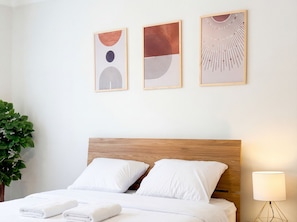 Een net opgemaakt bed met witte lakens en kussens, een houten hoofdbord en drie abstracte kunstwerken aan de muur.